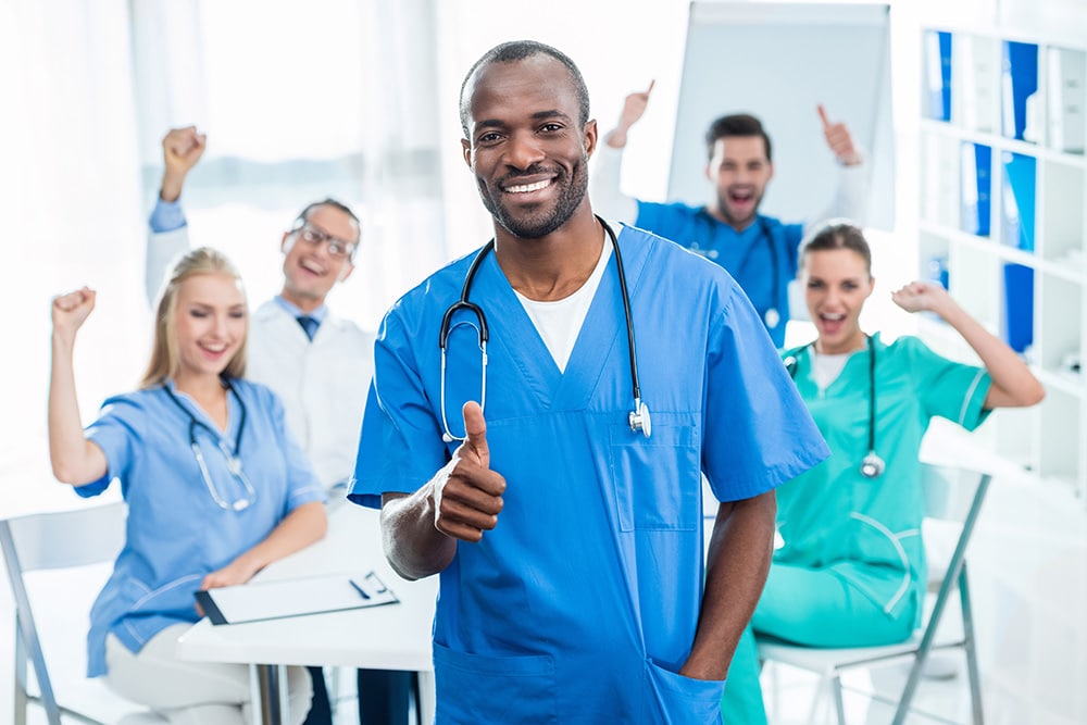 handsome nurse showing thumb up with colleagues sitting at table on background